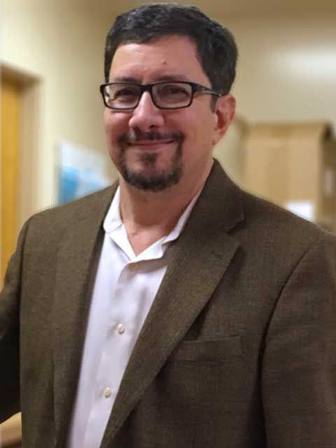 Man with glasses and goatee, wearing a brown blazer and white shirt, smiling.