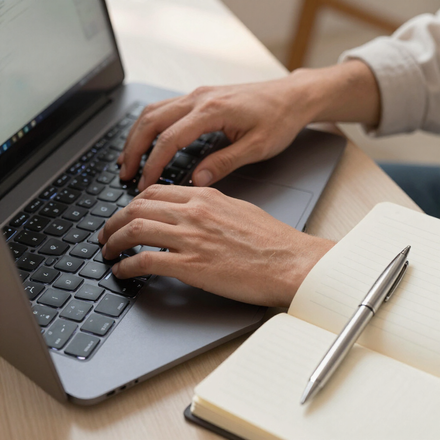 Handen typen op een laptop naast een notitieboekje en pen op een bureau.