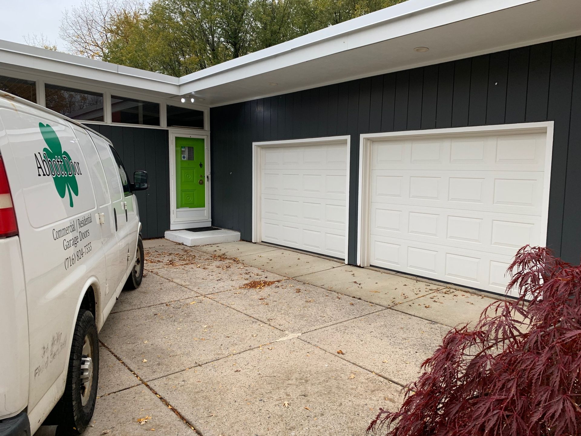 White van parked in front of a house with grey siding, two white garage doors, and a green front door.
