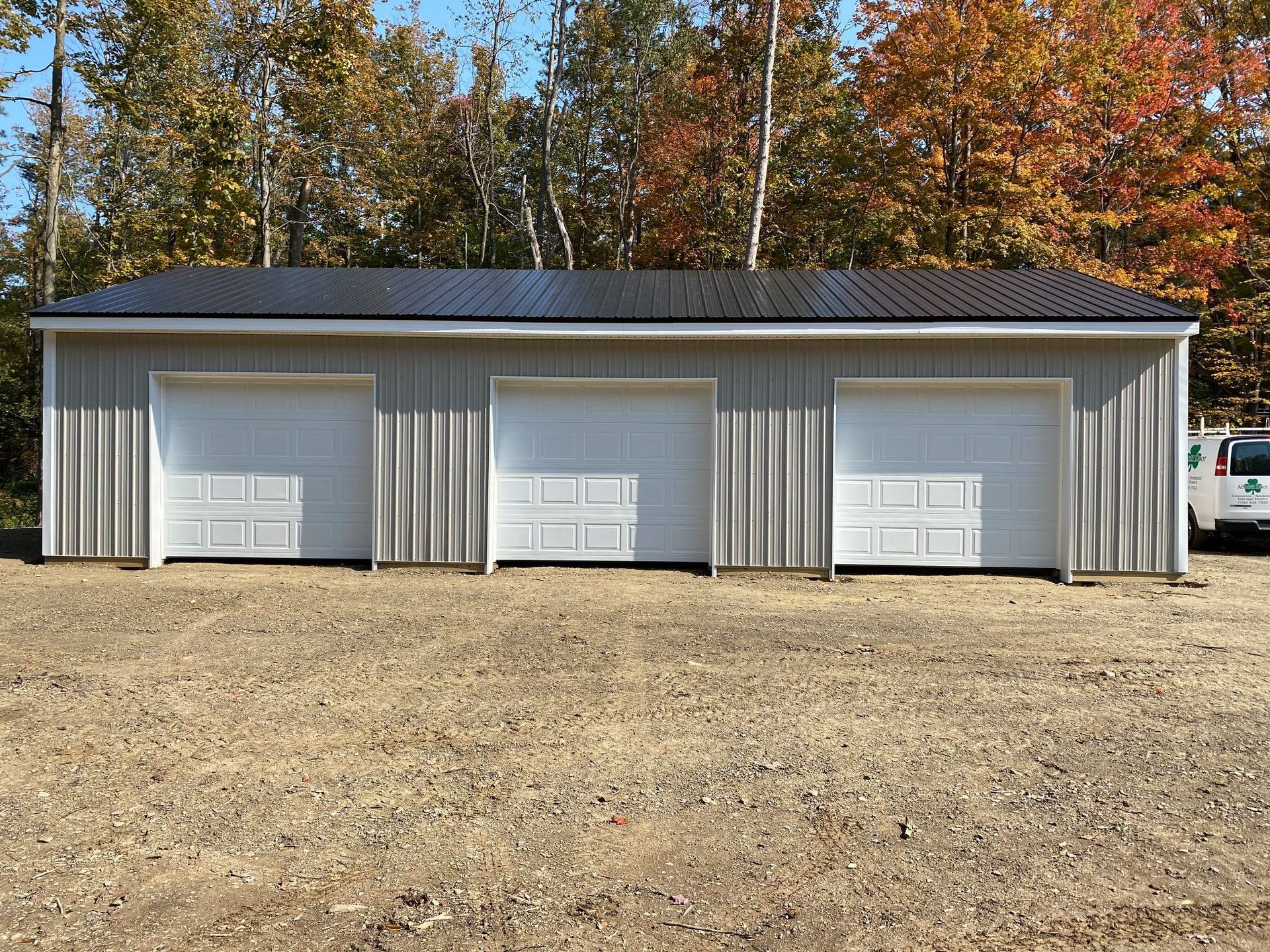A light gray three-car garage with white doors under a black metal roof. Forest in the background.