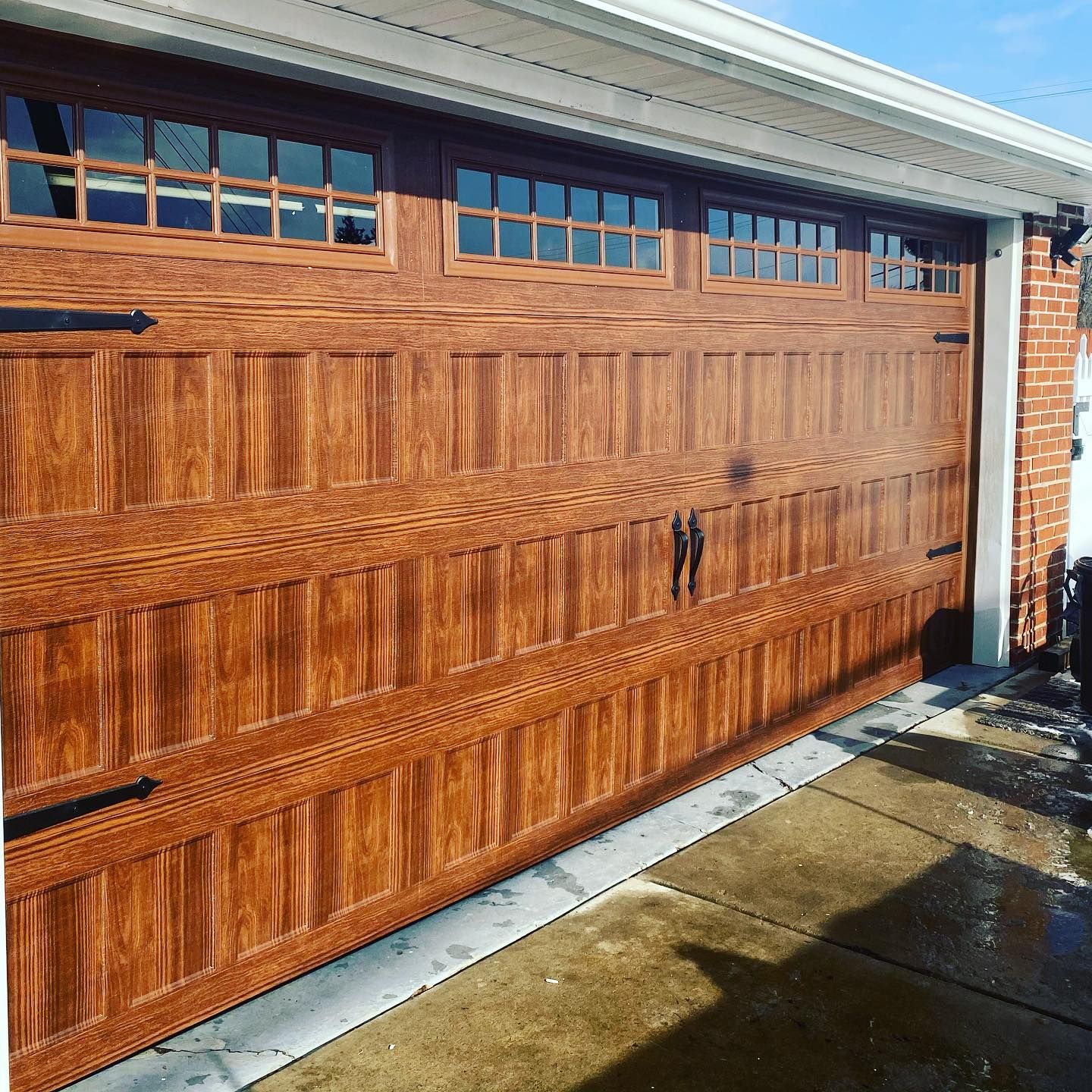 A brown wooden garage door with small windows at the top, black handles, and a brick wall on the right.