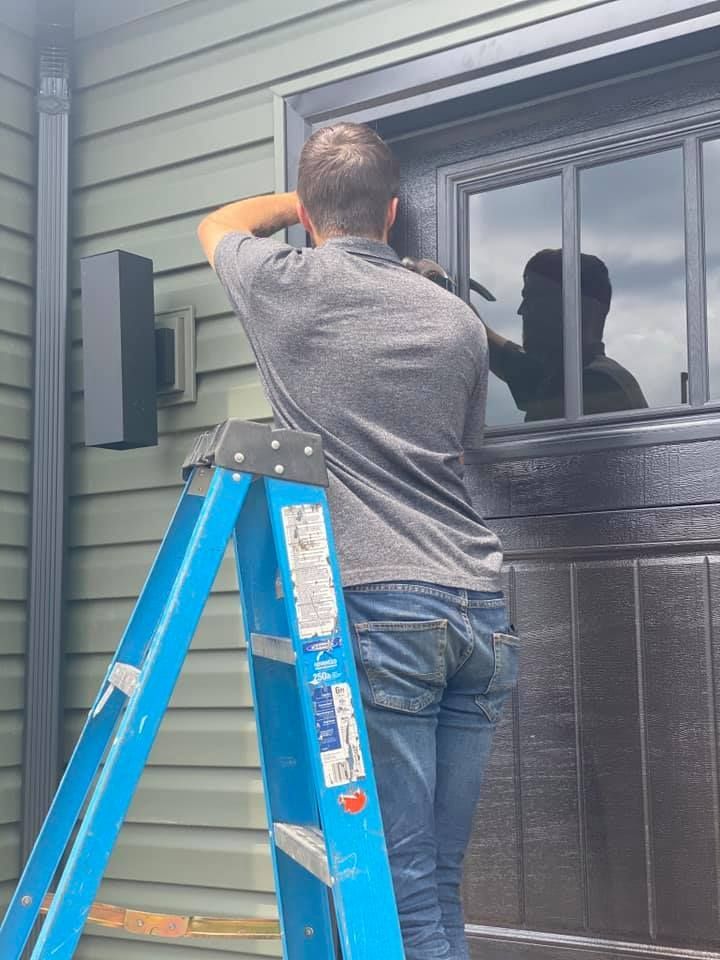 Man on a ladder installing something near a dark window, with green siding and a speaker nearby.