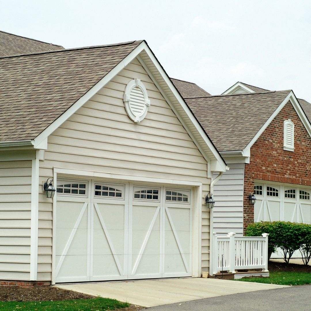 White garage doors with decorative trim on a beige building and brick structure with brown roof.
