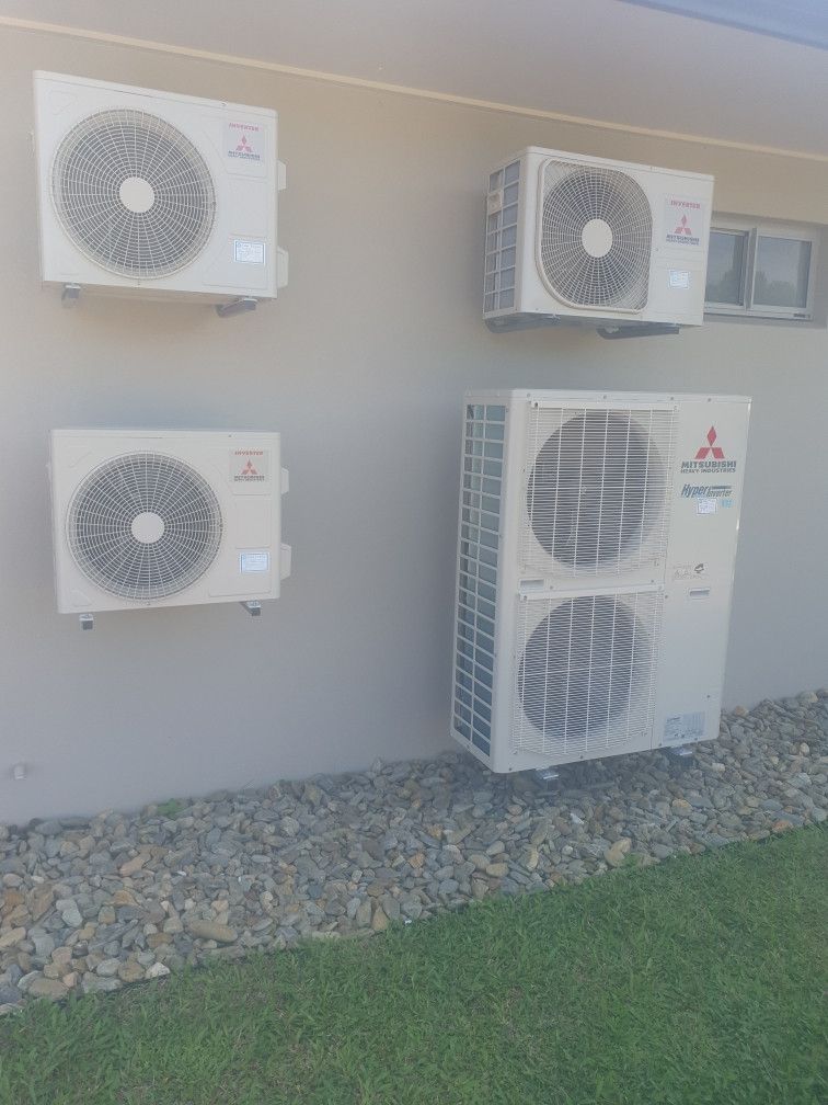 Four Mitsubishi air conditioning units mounted on a light beige wall next to gravel and grass.— Cool Tropics Air Conditioning & Refrigeration in Tablelands, QLD