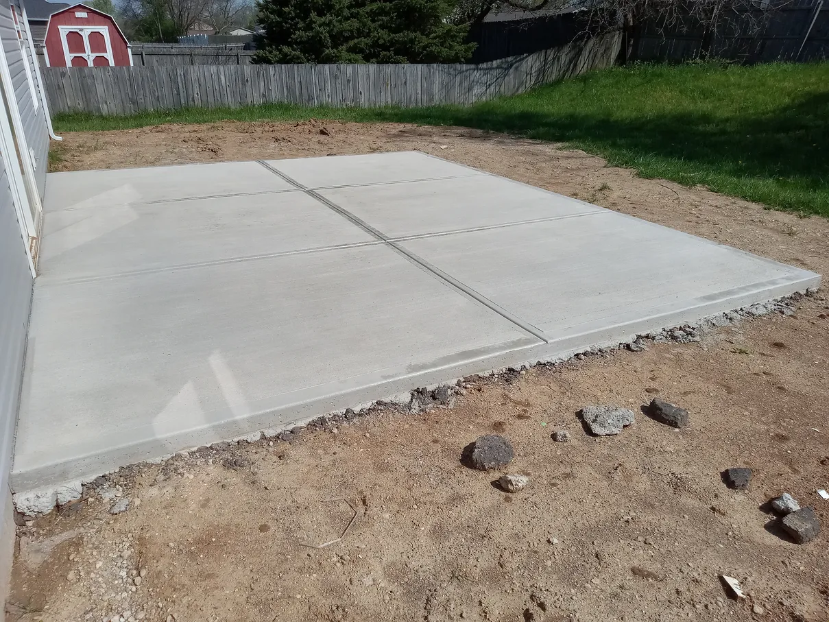 Newly poured concrete patio in a backyard, surrounded by dirt and grass, near a fence.
