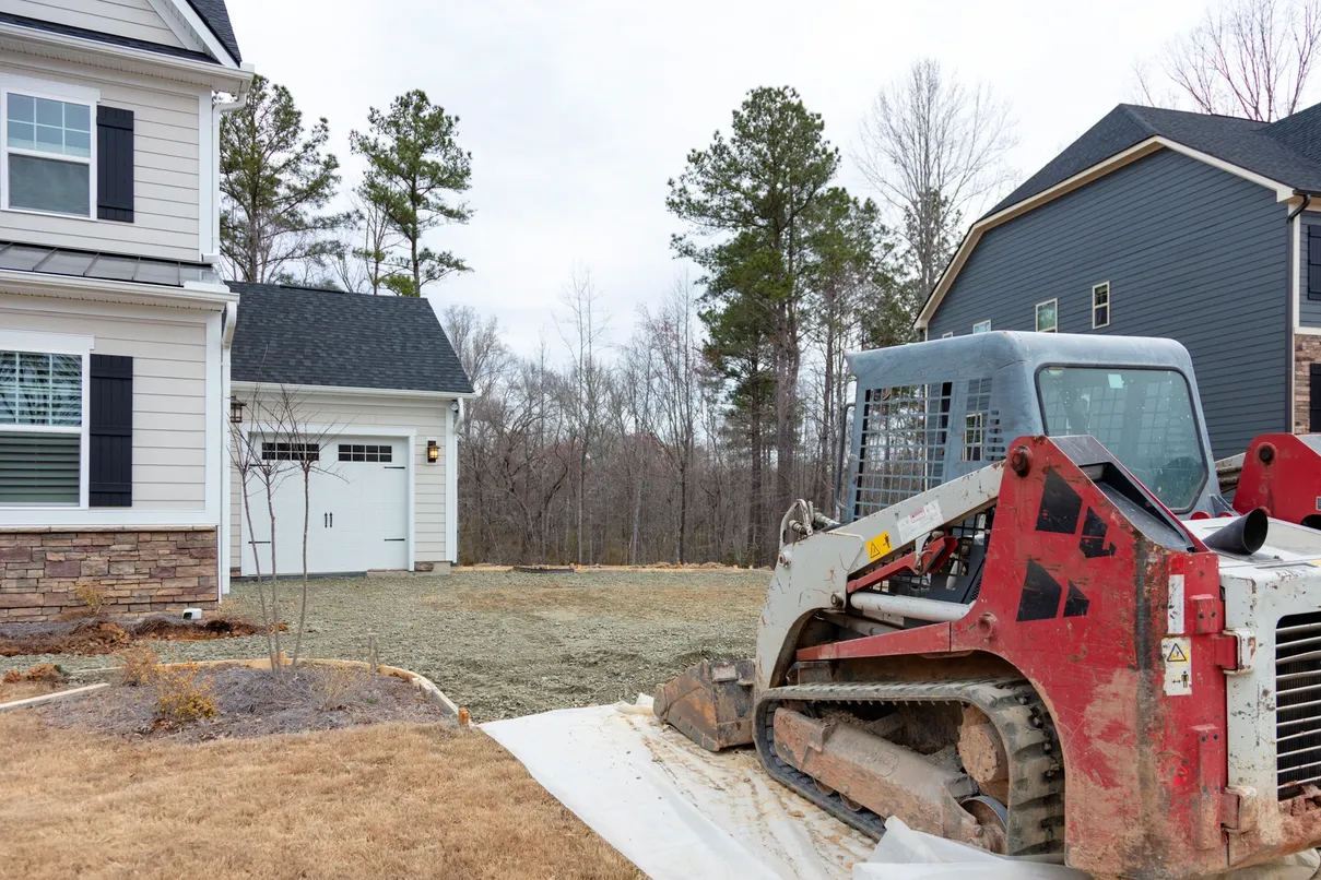 Skid steer next to a house, grading a gravel yard in front of a detached garage and trees.