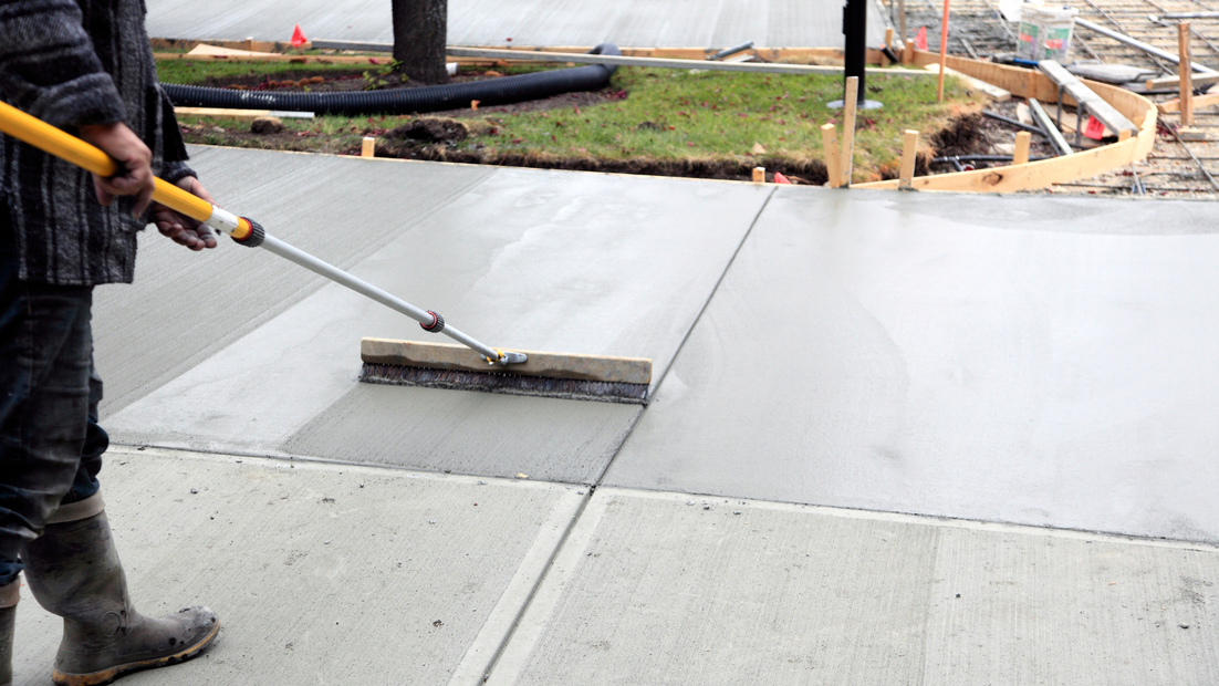 Person brushing wet concrete sidewalk with long-handled tool.