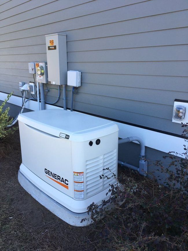 A Generac home backup generator, beige and white, installed outside a gray-sided house. Electrical panel visible above.