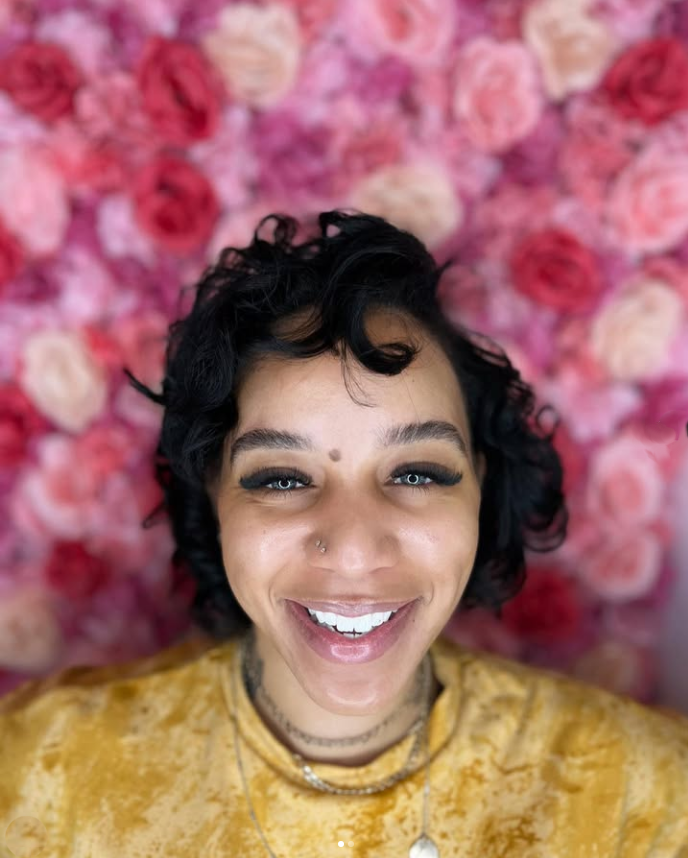 A woman is smiling in front of a wall of pink roses.