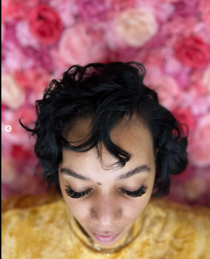A woman with short hair is laying in front of a wall of pink flowers.
