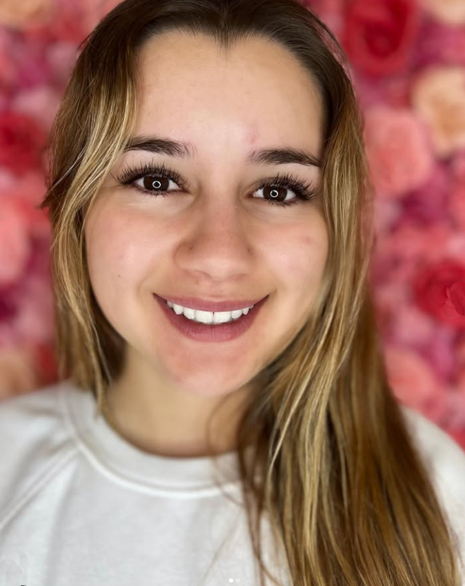 A woman is smiling in front of a wall of flowers.