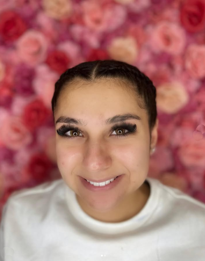 A woman is smiling in front of a wall of pink flowers.