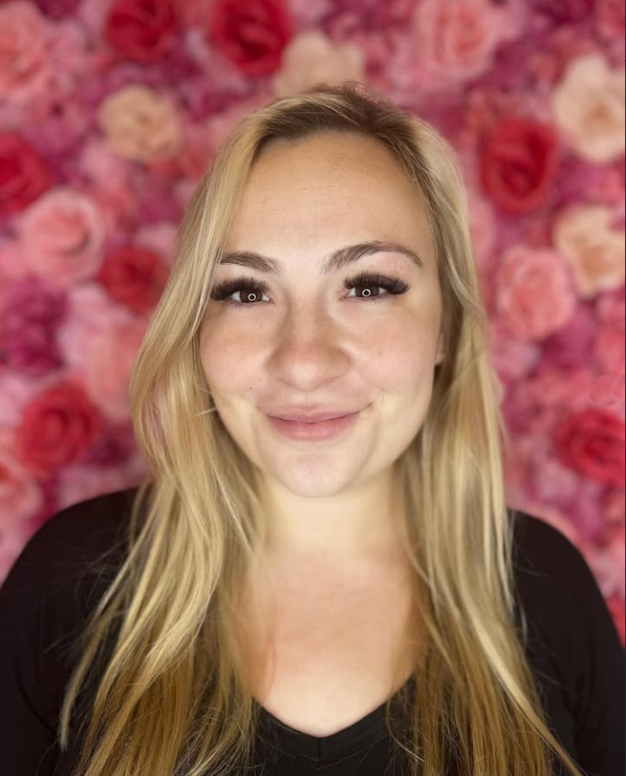 A woman is smiling in front of a wall of pink roses.