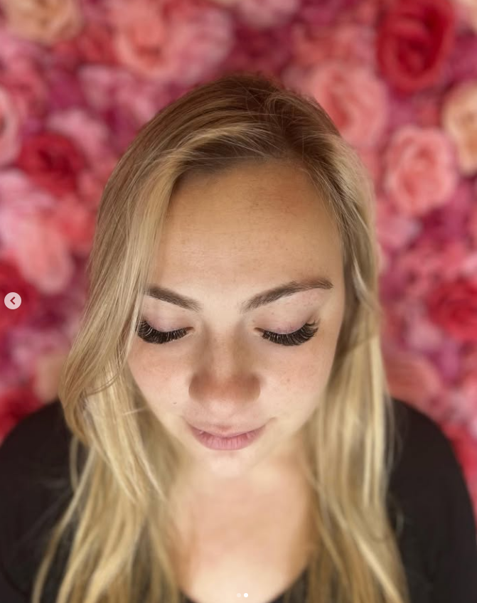 A woman with long blonde hair is looking down in front of a wall of pink roses.