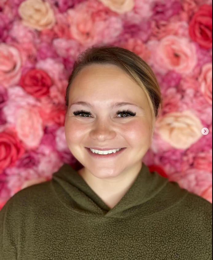 A woman is smiling in front of a wall of pink roses.