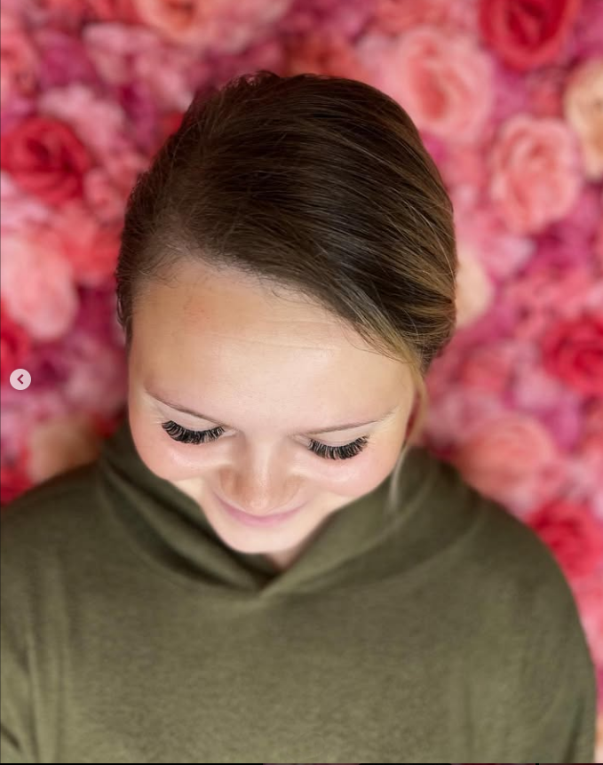 A woman with long eyelashes is standing in front of a wall of pink roses.