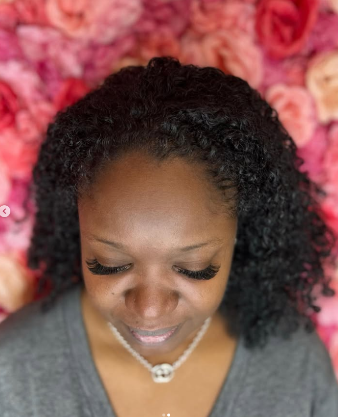 A woman with curly hair and a necklace is smiling in front of a wall of pink roses.