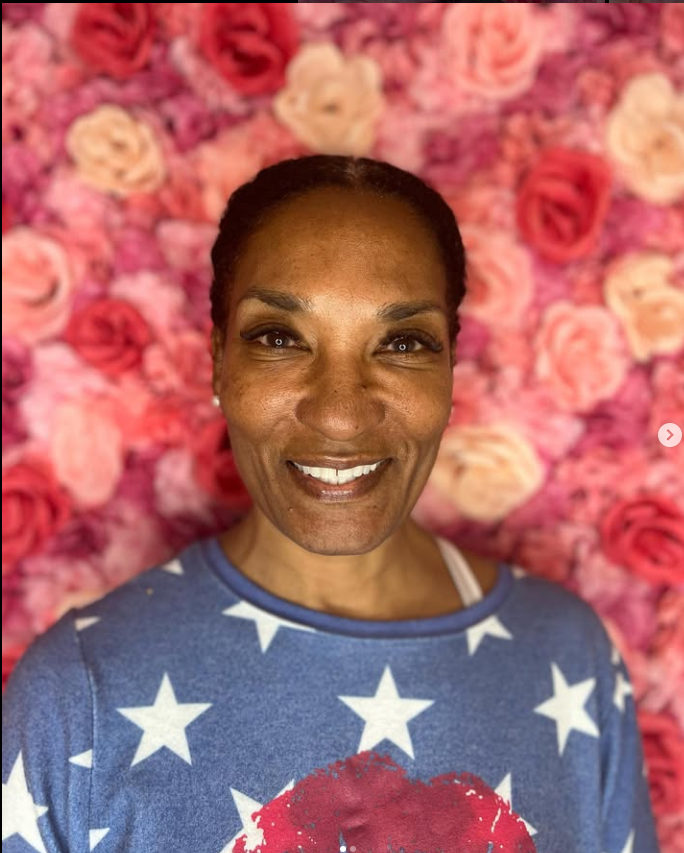 A woman is smiling in front of a wall of pink roses