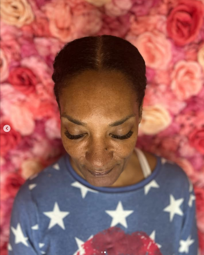 A woman in a blue shirt with white stars is standing in front of a wall of pink roses.