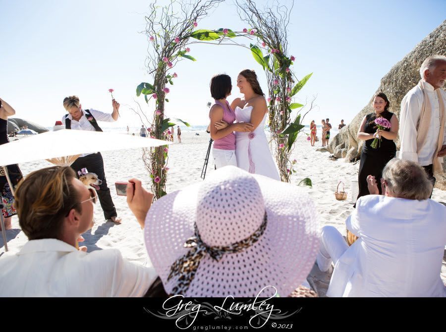 A woman in a white hat is taking a picture of a wedding ceremony on the beach