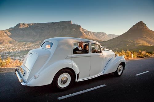 A white car is driving down a road with mountains in the background.