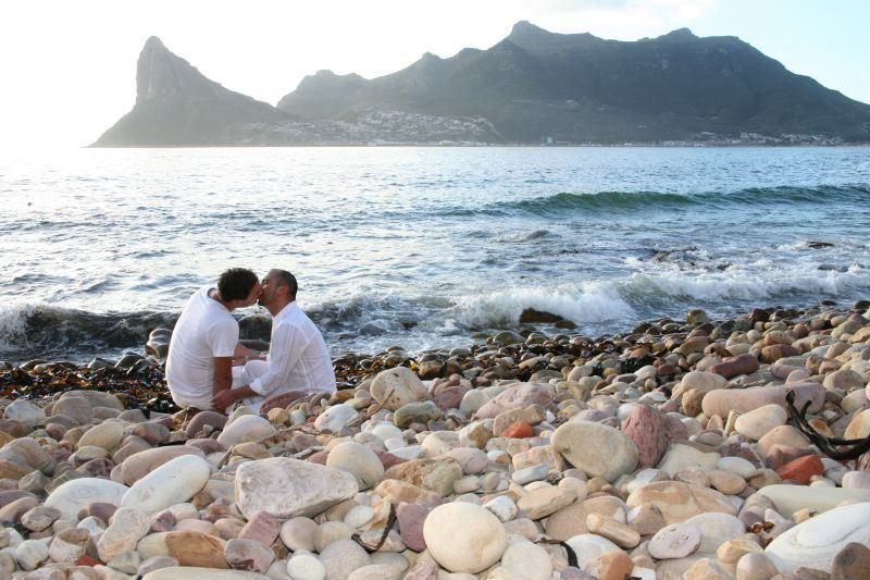 Two men kissing on a rocky beach with mountains in the background
