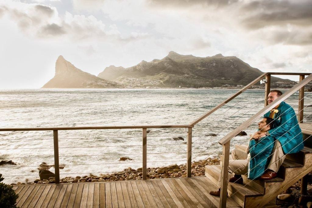 A couple is sitting on a wooden deck overlooking the ocean.