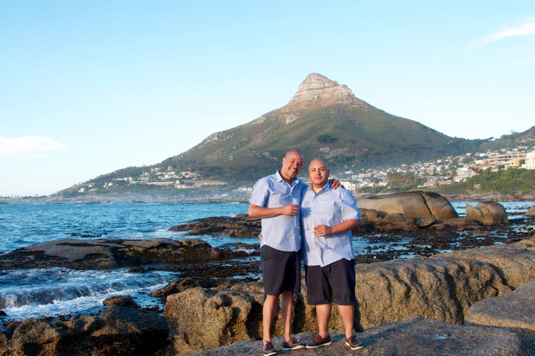 Two men are standing on a rocky beach with a mountain in the background.