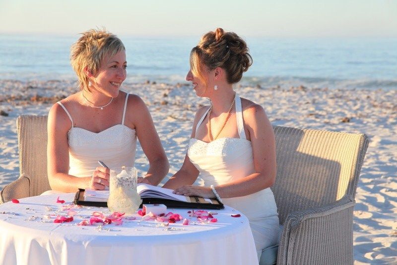 Two women in white dresses are sitting at a table on the beach.
