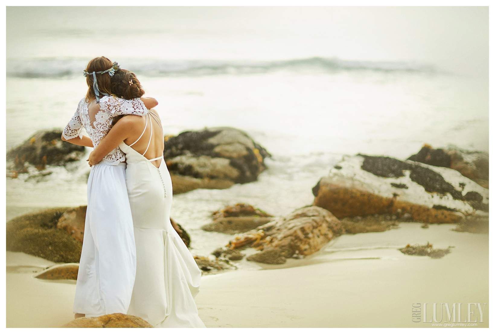 Two women in wedding dresses are hugging each other on the beach.