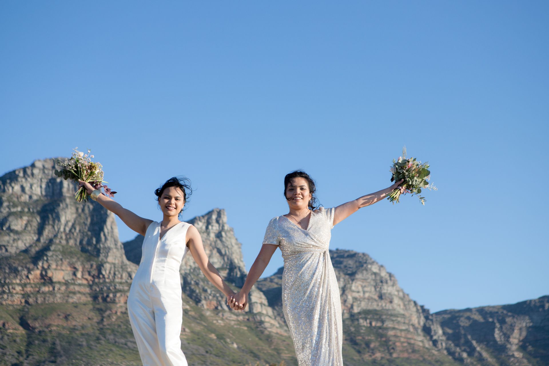 Two women in white dresses are holding hands in front of a mountain.