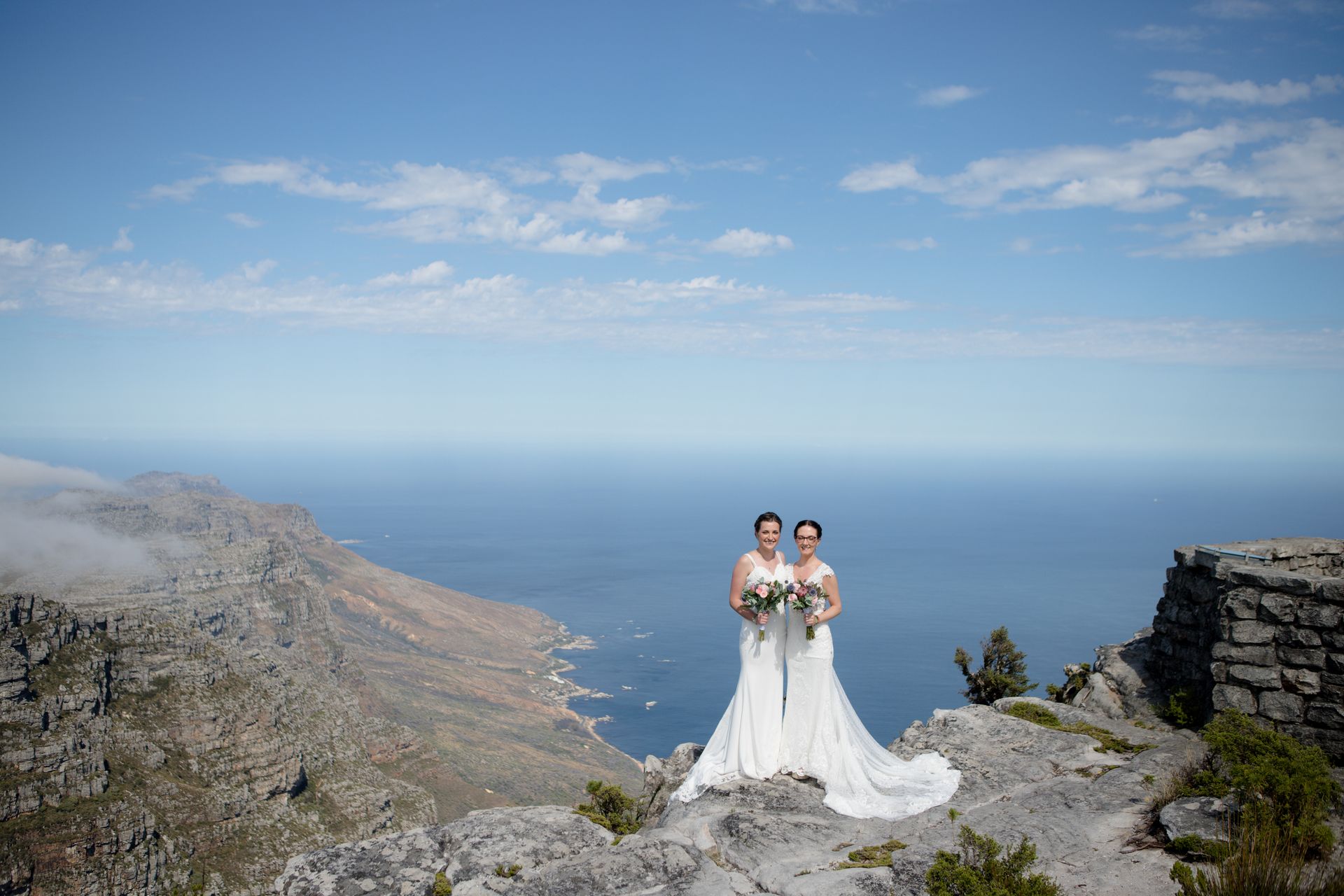 Two women in wedding dresses are standing on top of a mountain overlooking the ocean.
