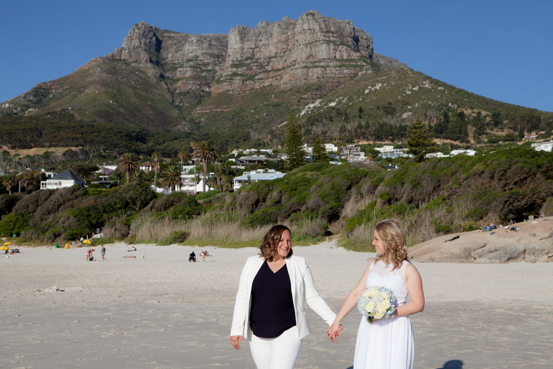 Two women are holding hands on a beach with a mountain in the background.