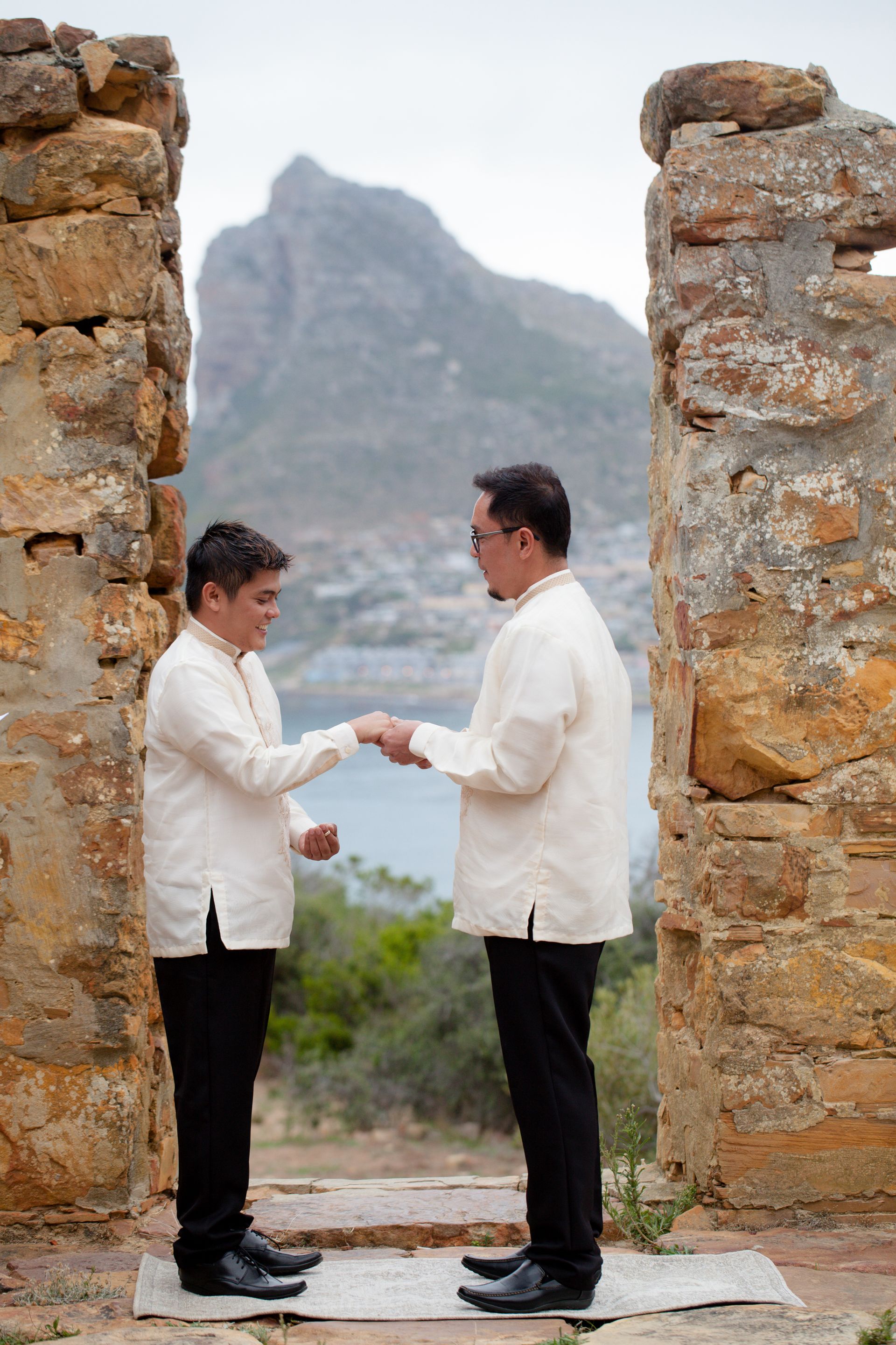 Two men are standing next to each other in front of a stone wall holding hands.