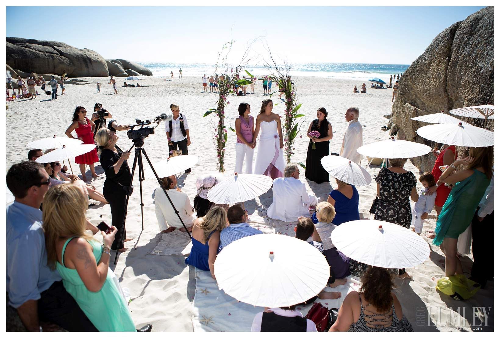 A group of people are sitting on the beach watching a wedding ceremony.