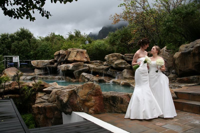 Two women in wedding dresses standing next to each other in front of a waterfall