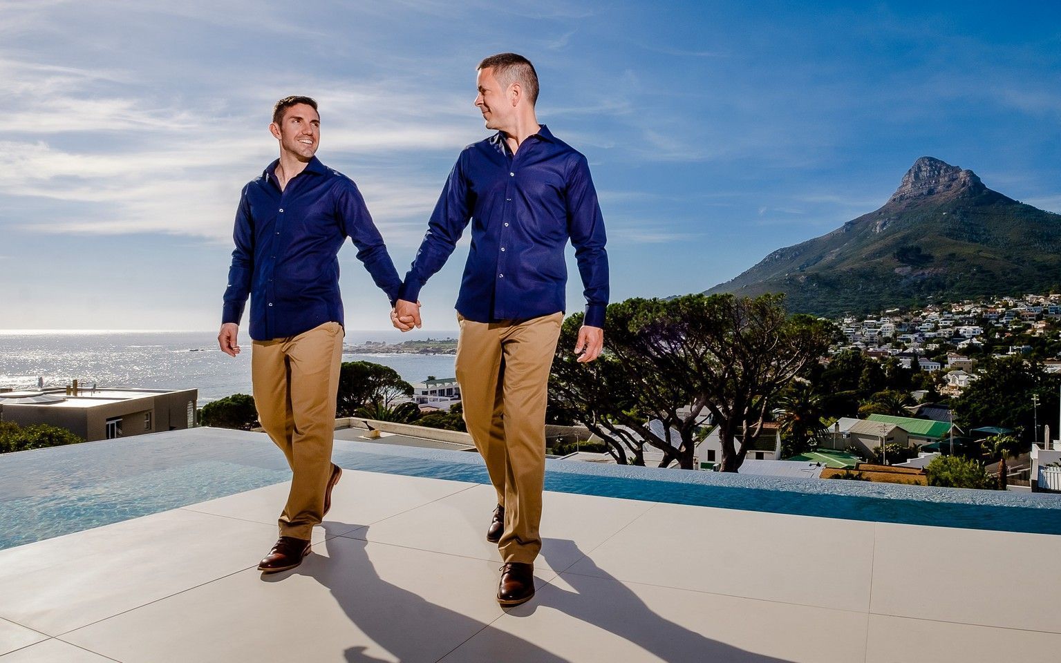 Two men are holding hands while walking on a balcony overlooking the ocean.