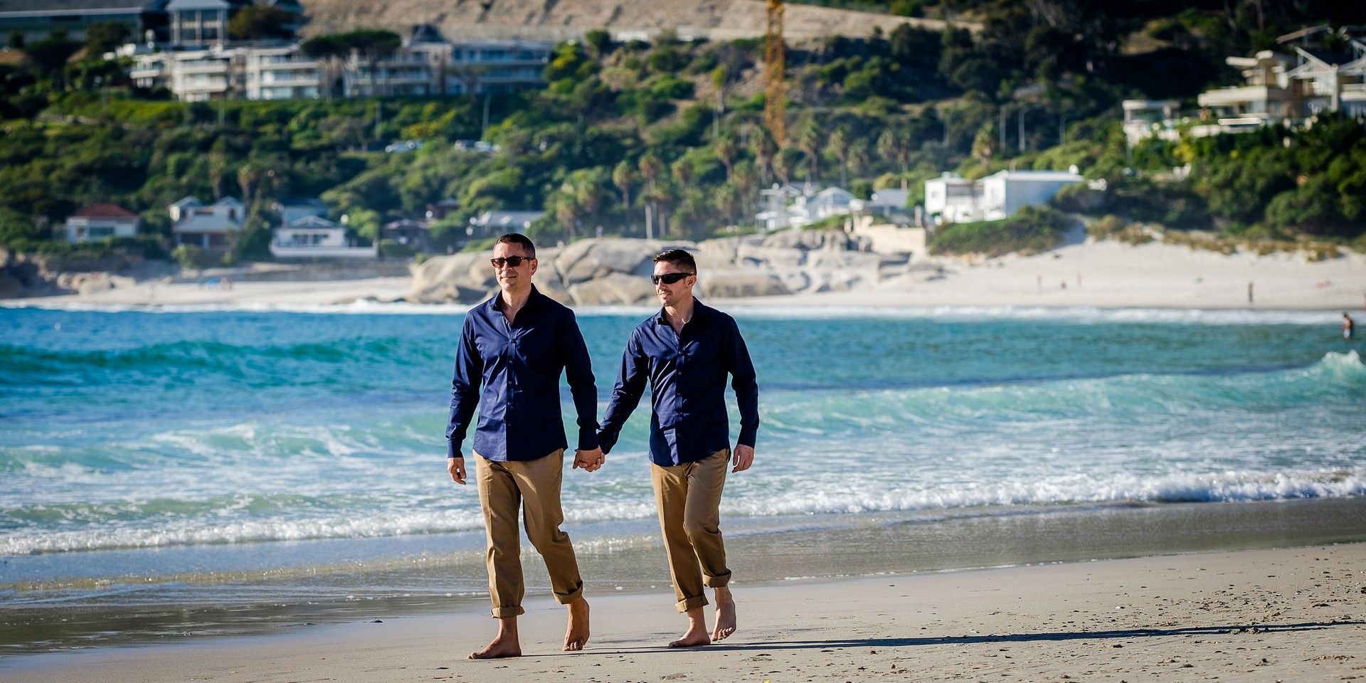 Two men are walking on the beach holding hands.