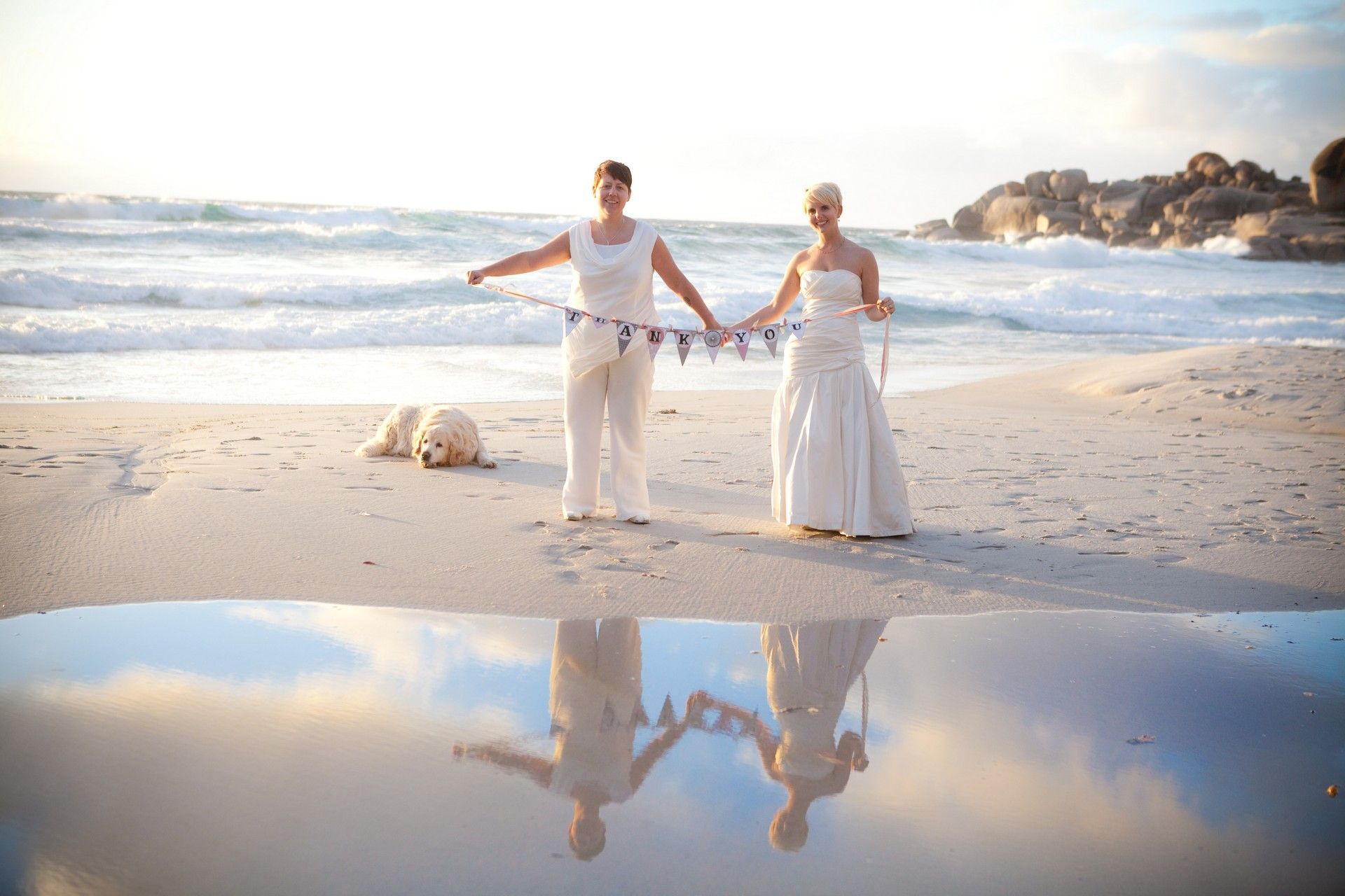 Two women in white dresses are standing on a beach holding hands.