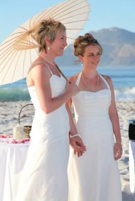 Two women in white dresses holding umbrellas on the beach