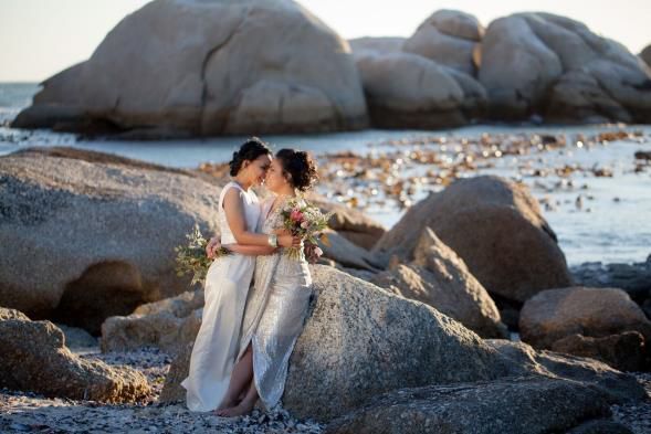 Two women are kissing on a rock on the beach.