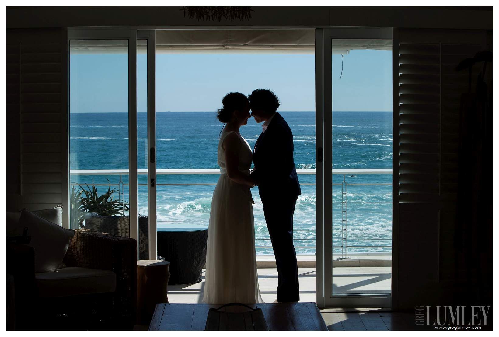 A bride and groom are kissing in front of a window overlooking the ocean.