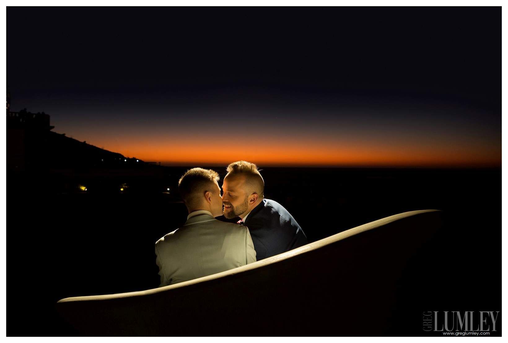 Two men are kissing on a bench at sunset.