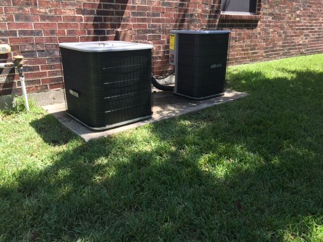 Two air conditioners are sitting in the grass in front of a brick building.