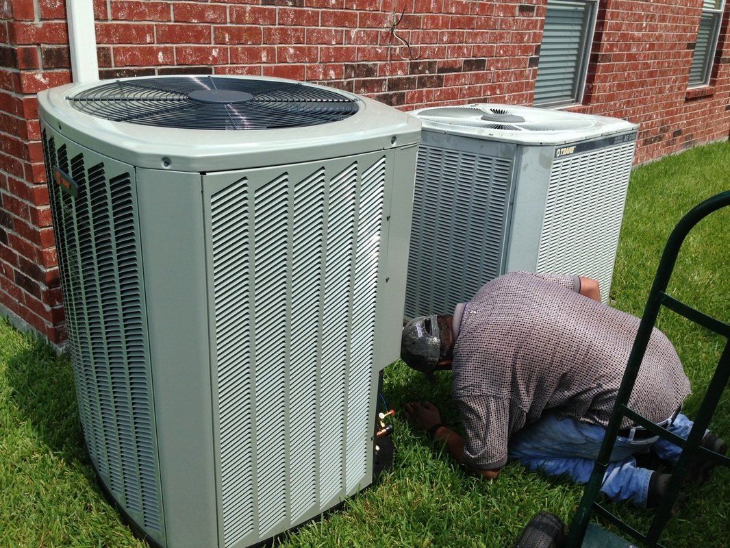 A man is kneeling in the grass next to two air conditioners.