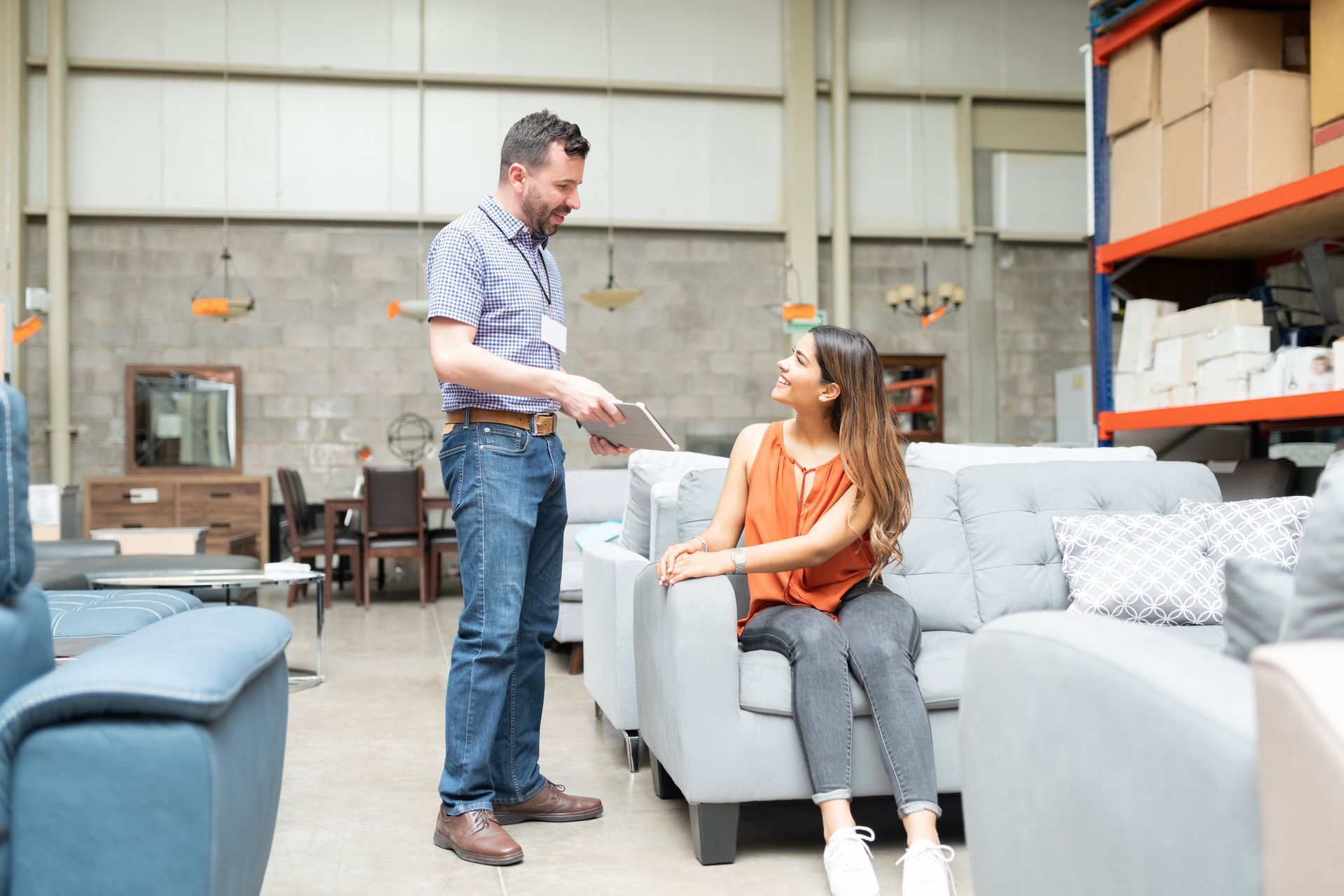 A male furniture salesman speaks to a woman sitting on a sofa inside a store.