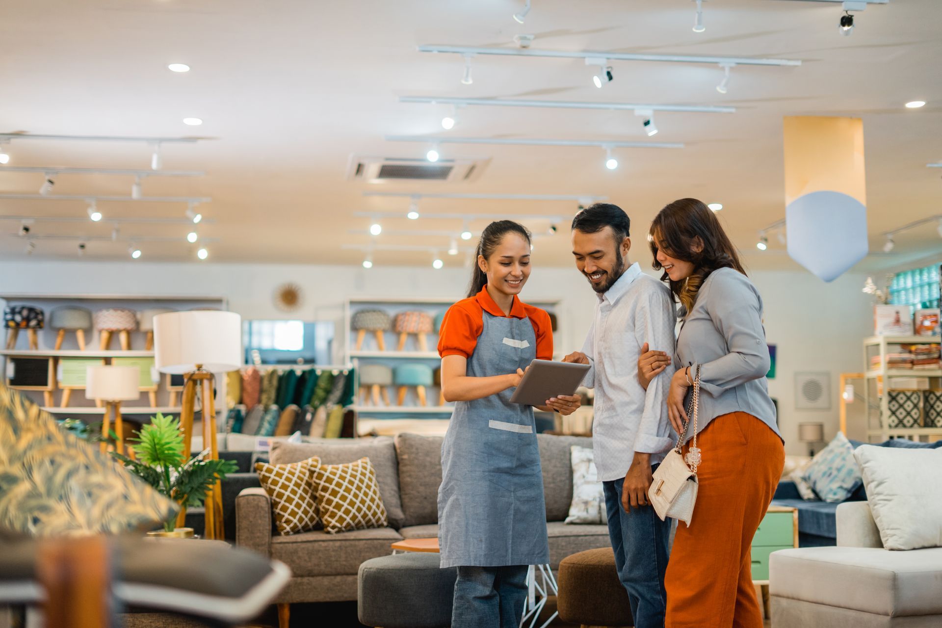 A store employee in a blue apron shows a tablet to a couple in a furniture showroom.