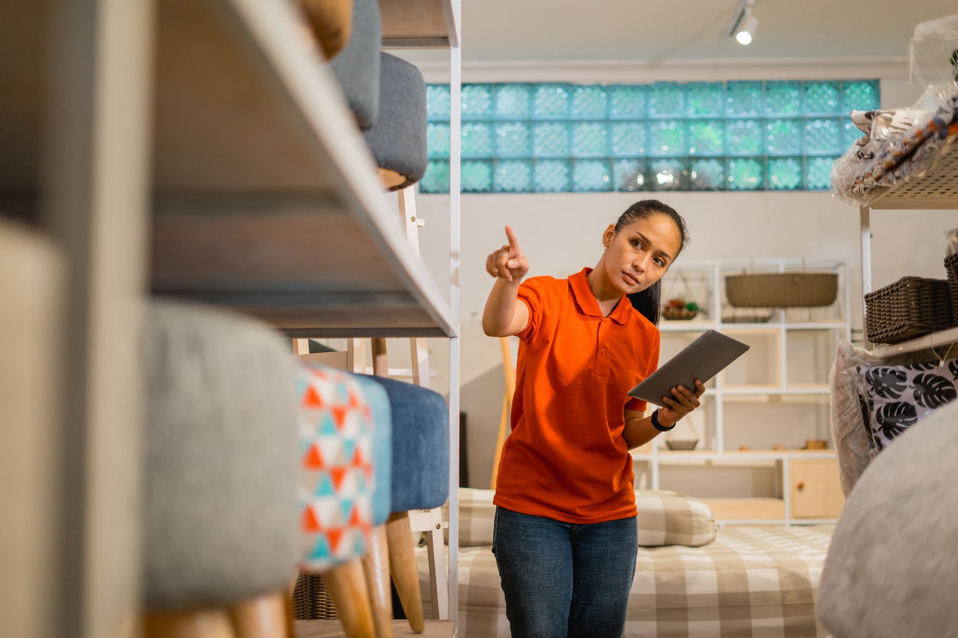 Woman in orange shirt, holding a tablet, pointing in a store with shelves of furniture.