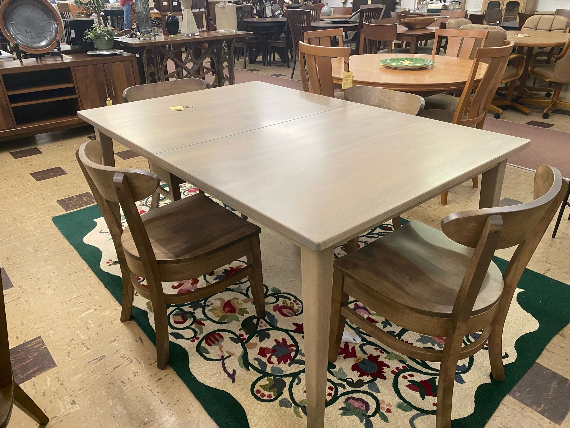 Wooden dining table with four chairs on patterned rug in a furniture store.