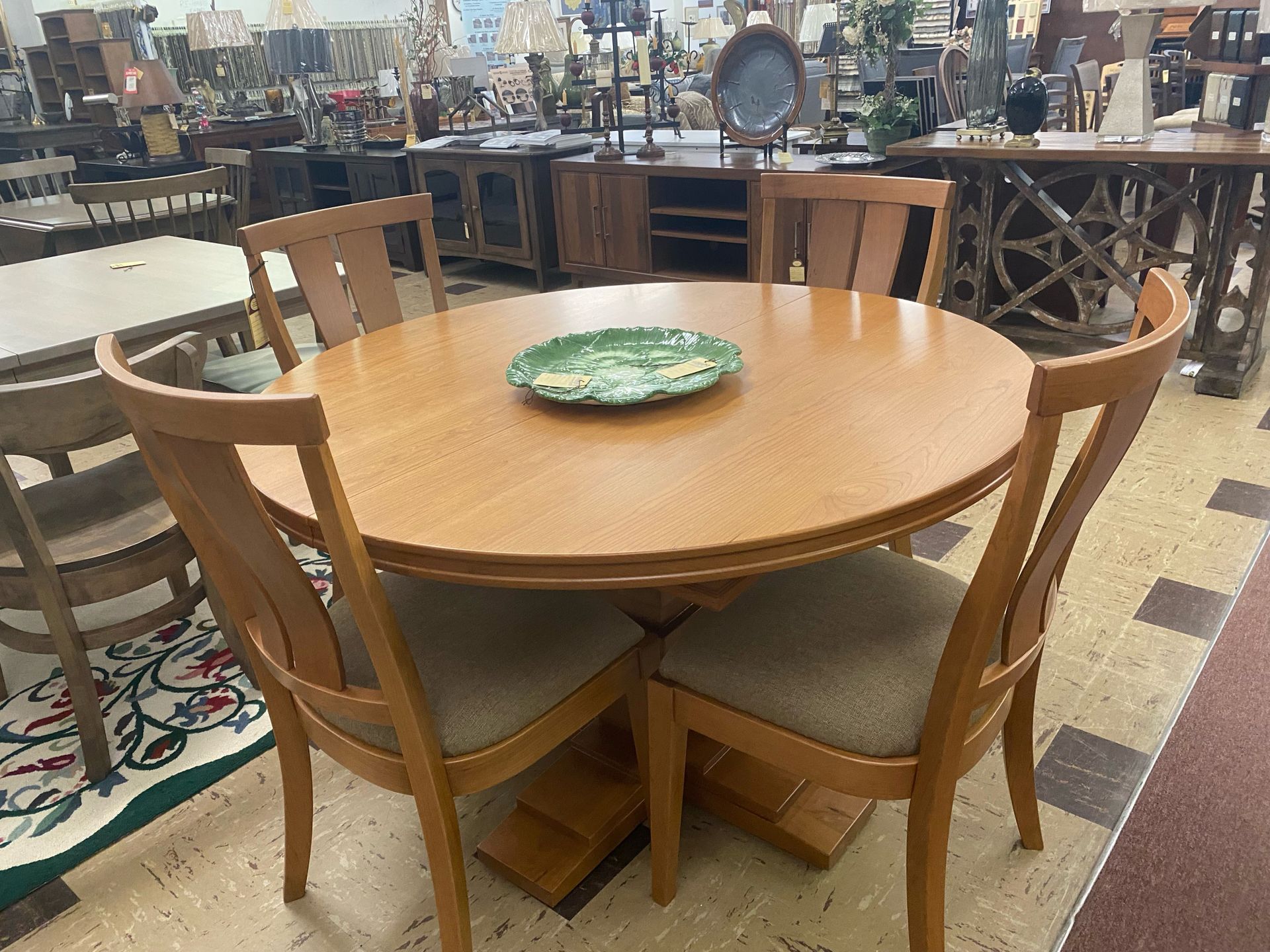 A wooden dining table with four chairs in a store. A decorative plate sits in the center.